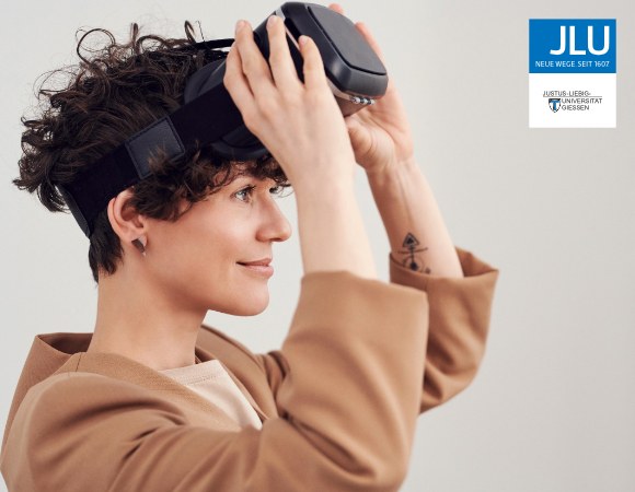 Ziergrafik: Eine Frau mit kurzen, gelockten, braunen Haaren und in einem beigen Anzug setzt eine Virtual Reality Brille auf und lächelt.